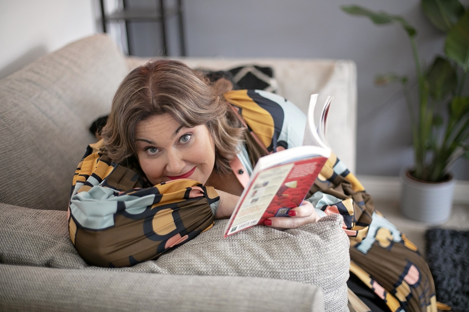 Meg Thompson reclining on a sofa with a book and looking towards the camera